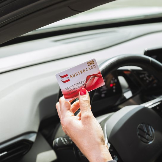 Hand hält eine AUSTROCARD im Innenraum eines Autos vor dem Lenkrad.